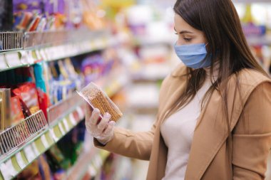 Koruyucu maskeli ve eldivenli bir kadının süpermarkette dikilirken mallara fiyat etiketi okuduğu yatay bir fotoğraf. Güzel genç kız markette alışveriş yaparken yılan seçiyor.