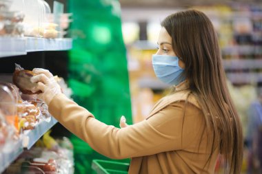 Koruyucu maskeli ve eldivenli bir kadın süpermarkette dururken fiyatlı etiketler okuyor. Güzel genç kız markette alışveriş yaparken yılan seçiyor. Alışveriş sırasında.