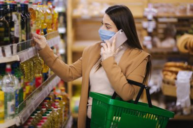 Süpermarkette koruyucu maske ve eldiven takan bir alıcı. Covid-19 pandemik karantinası sırasında alışveriş. Çürümez zekâ, ev eşyaları için kiler hazırlığı satın aldı. Telefonla kadın mızrağı