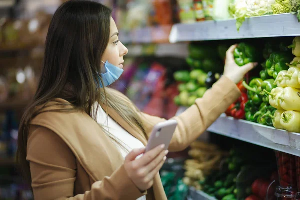 Happy woman in face mask hold phone and choose vegetables in supermarket. Beautiful young business woman choosing groceries by stand with vegetables. Quarantine. Covid-19