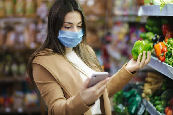 戴面具的快乐女人拿着电话，在超市里挑选蔬菜。漂亮的年轻女商人站在蔬菜旁边挑选食品杂货。隔离。Covid-19 — 图库照片
