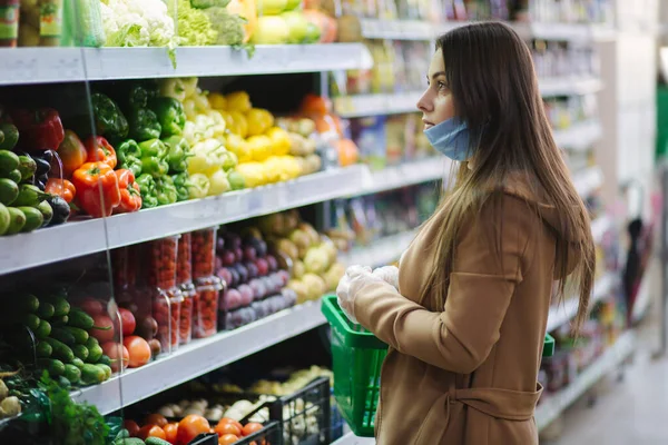 Ευτυχισμένη γυναίκα με προστατευτική μάσκα που παίρνει φρέσκα λαχανικά ενώ στέκεται δίπλα σε παντοπωλεία στο σούπερ μάρκετ. Όμορφη νεαρή κοπέλα με καλάθι τροφίμων επιλέγοντας το φαγητό από περίπτερο με λαχανικά. Χριστουγεννιάτικες αγορές — Φωτογραφία Αρχείου