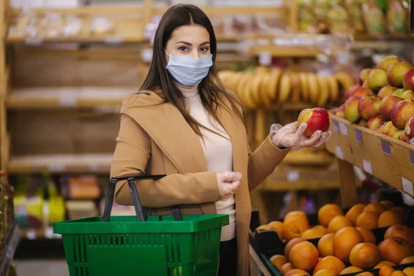 Νεαρή γυναίκα με προστατευτικά γάντια και μάσκα προσώπου κρατά όμορφο φρέσκο μήλο στο χέρι. Όμορφη νεαρή κοπέλα με καλάθι τροφίμων επιλογή τροφίμων από σταθεί με φρούτα. Ψώνια στην καραντίνα. Covid-19 — Φωτογραφία Αρχείου