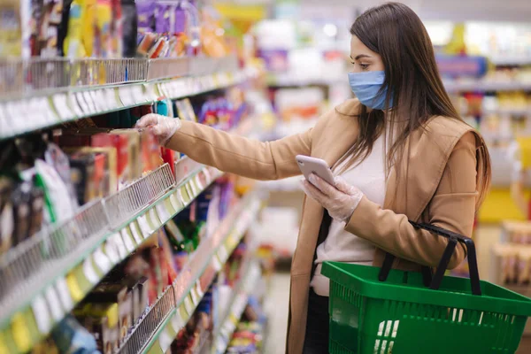 La mujer con máscara protectora y guantes escanea la etiqueta con el precio usando el teléfono en las mercancías mientras está de pie en el supermercado. Buscando en internet. Hermosa chica joven eligiendo snaks mientras hace compras en el supermercado — Foto de Stock