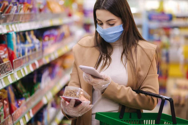 La mujer con máscara protectora y guantes escanea la etiqueta con el precio usando el teléfono en las mercancías mientras está de pie en el supermercado. Buscando en internet. Hermosa chica joven eligiendo snaks mientras hace compras en el supermercado — Foto de Stock