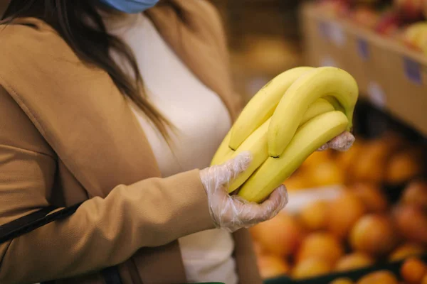 Закрытая молодая женщина в защитных перчатках и маске для лица держит в руках красивые свежие бананы. Красивая молодая девушка с корзиной продуктов питания, выбирая пищу стенд с фруктами. Покупки во время карантина — стоковое фото