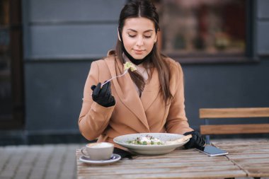 Terastaki kafede öğle yemeği yiyen güzel genç bir kadın. Siyah koruyucu maskeli bir kadın. Kadın yemek yerken maskesini çıkarıyor.