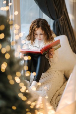 Güzel küçük kız Noel tatili boyunca pencere eşiğinde kitap okuyor.