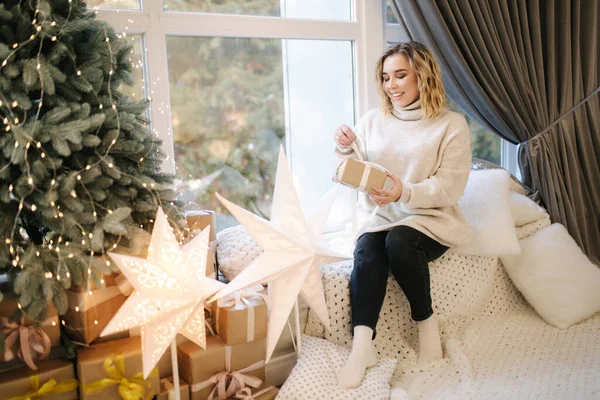 Blont hår kvinna i luvtröja öppna present på juletid. Elegant kvinna står vid en stor julgran framför fönstret — Stockfoto