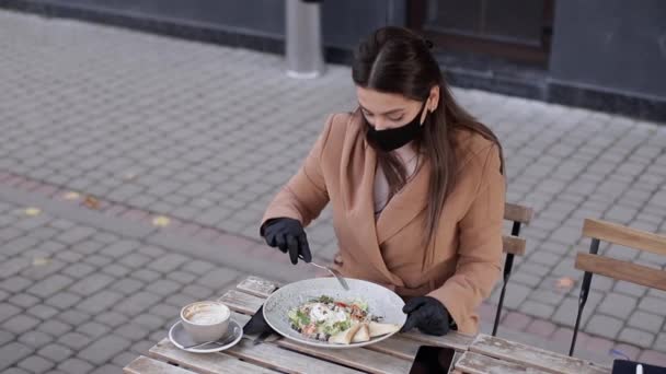 Karantänkafékoncept på terrassen. Attraktiv ung kvinna sitter på kafé på egen hand. Kvinnan äter under rasten. Kvinna i svart ansiktsmask och handskar äter vegansallad — Stockvideo