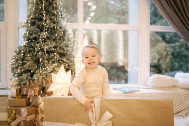 Sevimli küçük çocuk Noel ağacının ve büyük pencerenin önünde büyük bir hediye tutuyor.