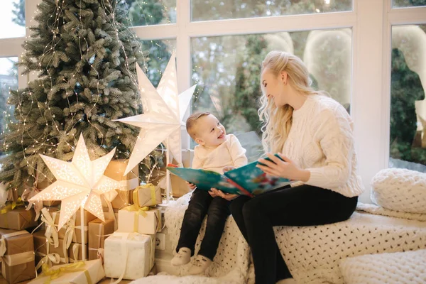 Boldog kisfiú könyvet olvas anyával az ablakpárkányon. Anya és az aranyos fia együtt töltenek egy kis időt a karácsonyi szünetben. Díszterem — Stock Fotó