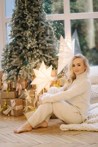 Vacker kvinna som sitter på fönsterbrädan vid julgranen. Blont hår kvinna i vit kostym har vila hemma. Julstämning — Stockfoto