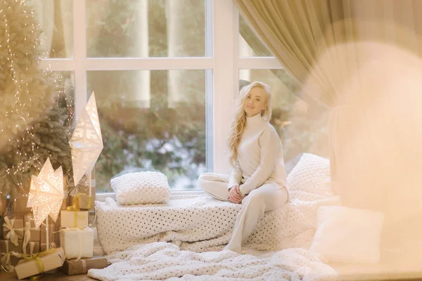 Piękna kobieta siedząca na parapecie przy choince. Blondynka w białym kostiumie odpoczywa w domu. Świąteczny nastrój — Zdjęcie stockowe