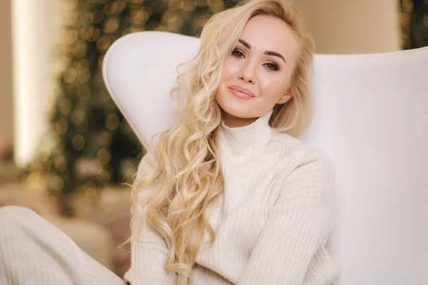 Atraktivní mladá blond žena v bílém pleteném obleku sedí na bílém křesle před vánočním stromečkem. Zimní dovolená — Stock fotografie
