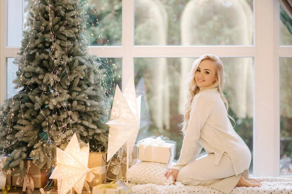 Gyönyörű nő ül az ablakpárkányon a karácsonyfa mellett. A szőke, fehér ruhás nő otthon pihen. Karácsonyi hangulat — Stock Fotó