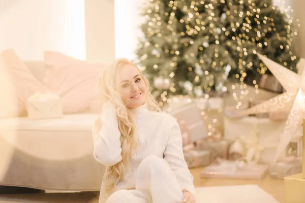 Blont hår kvinna i vit stickad kostym sitter hemma vid julgranen. Vacker kvinna på semester, julstämning — Stockfoto