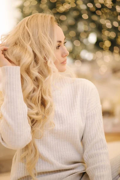 Portrait of Blond hair woman in white knitted suit sitting at home by Christmas tree. Beautiful woman at holiday, Christmas mood — Stock Photo, Image