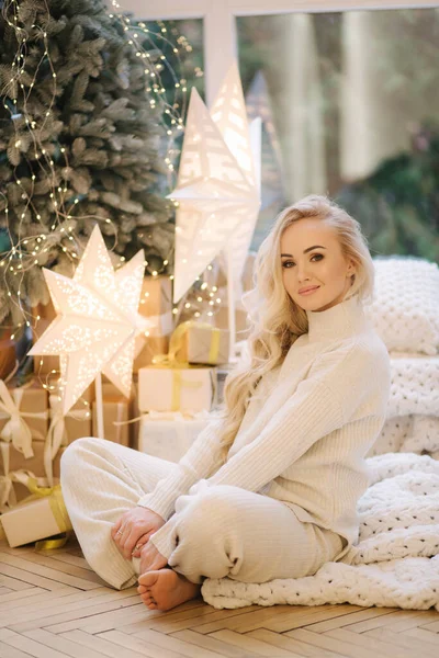 Blont hår kvinna i vit stickad kostym sitter hemma vid julgranen. Vacker kvinna på vinterlov, julstämning — Stockfoto