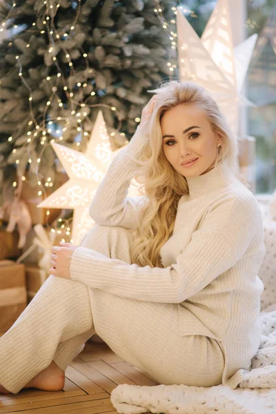 Blondynka w białym garniturze siedząca w domu przy choince. Piękna kobieta na feriach zimowych, świąteczny nastrój — Zdjęcie stockowe