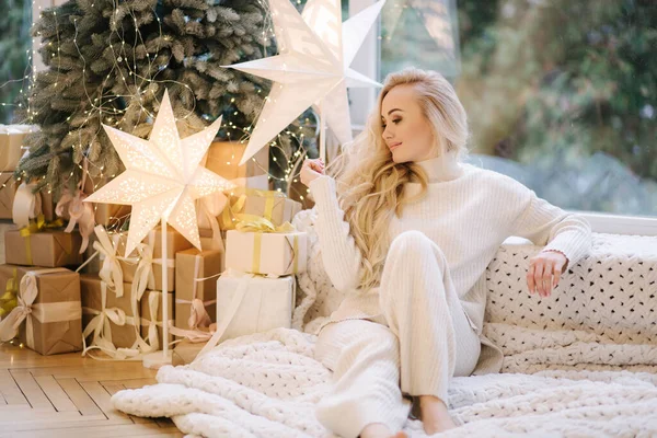 Beautiful woman sitting on windowsill by Christmas tree. Blond hair woman in white costume have rest at home. Christmas mood — Stock Photo, Image