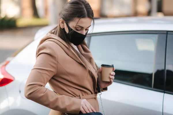 Woman in black face mask hold cuf of coffee by the car and look car key. Woman open car. Protectiv mask in quarantine. Cov-19