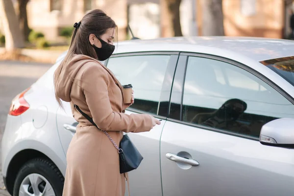 Woman in black face mask hold cuf of coffee by the car and look car key. Woman open car. Protectiv mask in quarantine. Cov-19