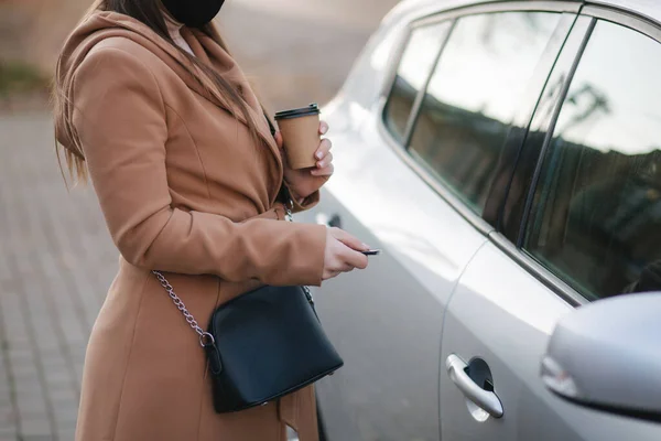 stock image Woman in black face mask hold cuf of coffee by the car and look car key. Woman open car. Protectiv mask in quarantine. Cov-19