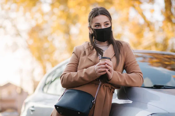 Beautiful young woman in black face mask stand by the car and drink coffee. Happy young woman have a break. Female pulled off mask for drinking cappucino outdoors