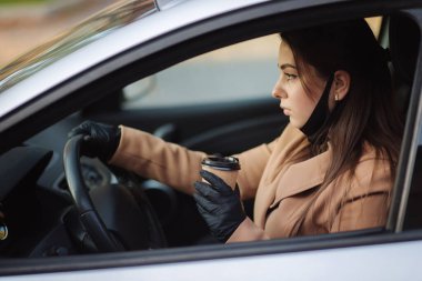 Yüz maskeli bir kadın Coronavirus salgını sırasında arabasını bir fincan kahveyle kullanıyor, Covid-19. Cappucino içmek için koruyucu maskeyi çıkarmış bir kadın.