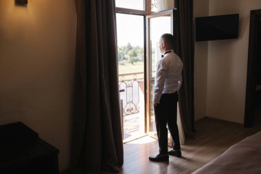 Damat, otelin balkonunun yanındaki pencerede duruyor. Yakışıklı damat gelini bekliyor.