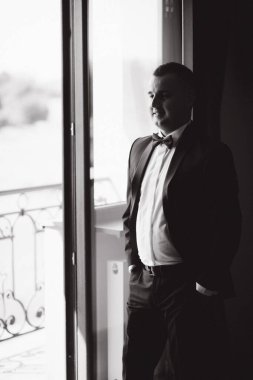Damat, otelin balkonunun yanındaki pencerede duruyor. Yakışıklı damat gelini bekliyor.
