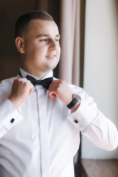 Handsome groom stand by window and straightens his tie bow