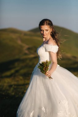 Dağdaki tepede duran genç ve güzel gelin. Dışarısı rüzgarlı. Arkaplanda yeşil tepeler