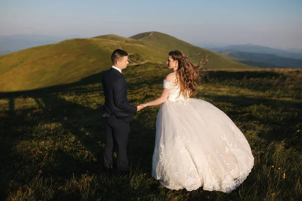 Γαμπρός και νύφη στο λόφο στα βουνά. Γάμου ζευγάρι μπροστά από το πράσινο λόφο. Νιόπαντροι απολαμβάνοντας ρομαντικές στιγμές στα βουνά στο ηλιοβασίλεμα στην όμορφη καλοκαιρινή μέρα — Φωτογραφία Αρχείου
