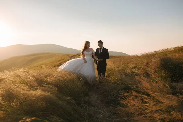 Ganzkörperporträt der attraktiven Braut und des stilvollen Bräutigams, die romantische Momente in den Bergen bei Sonnenuntergang an einem schönen Sommertag genießen. Hochzeitspaar im Feld — Stockfoto
