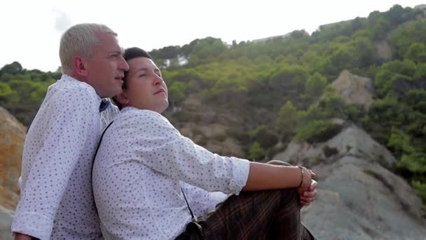 Två glada man med söt hund sitter på stenar längs stranden och pratar, Happy gay par kommunikation. Turister till havs — Stockvideo