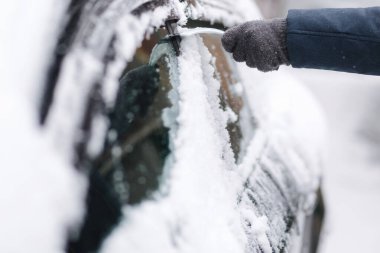 Bir adam kar kazıyıcı ile bir arabanın karlı camını temizliyor. Kazıyıcıya odaklan. Soğuk, karlı ve soğuk bir sabah. Siyah araba.