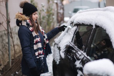Bir kadın kar kazıyıcı ile bir arabanın karlı camını temizliyor. Güzel, sıcacık giyimli, açık havada arabasını temizleyen bir kadın. Soğuk, karlı ve soğuk bir sabah. Siyah araba.