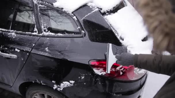 Młody człowiek czyszczący śnieg z przedniej szyby samochodu w pobliżu garażu w zimowy dzień — Wideo stockowe