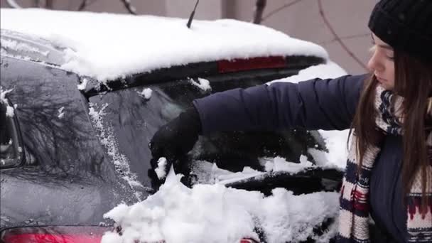 Жінка прибирає снігове вікно на машині зі сніговим скребком. Гарненька жінка воювала одягнена в чисту машину на відкритому повітрі. Холодний снігопад і морозний ранок. Чорний автомобіль — стокове відео
