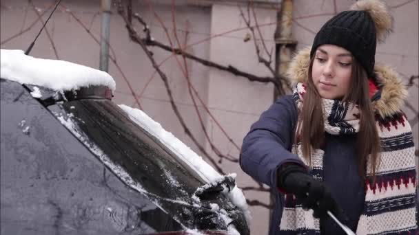 Жінка прибирає снігове вікно на машині зі сніговим скребком. Гарненька жінка воювала одягнена в чисту машину на відкритому повітрі. Холодний снігопад і морозний ранок. Чорний автомобіль — стокове відео