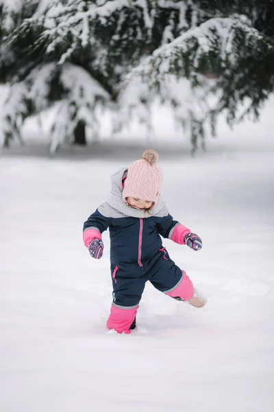 Glückliches kleines Mädchen, das im Park auf Schnee spaziert. Entzückendes kleines Mädchen mit rosa Hut und Overall, das an einem verschneiten Wintertag im Wald spaziert — Stockfoto