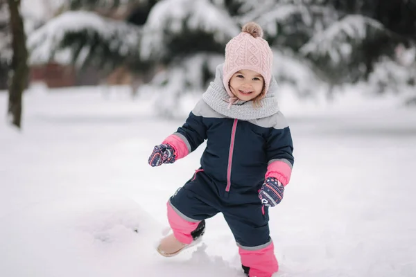 Ragazzina felice che cammina nel parco sulla neve. Adorabile bambina con cappello rosa e tuta da lavoro che cammina nei boschi in una giornata invernale innevata — Foto Stock
