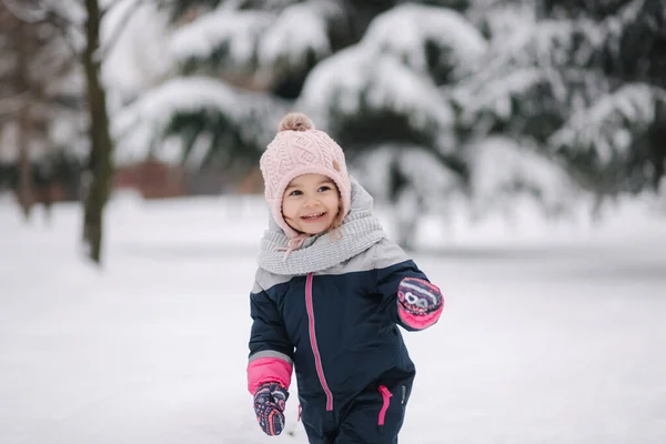 Menina feliz andando no parque na neve. Menina adorável em um chapéu rosa e macacão andando na floresta em um dia nevado de inverno — Fotografia de Stock