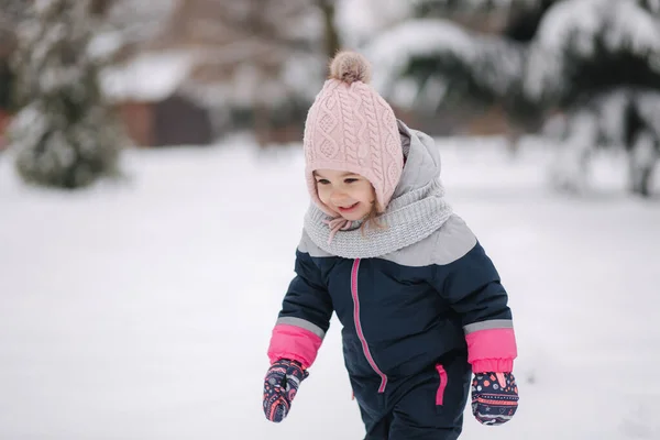 Gelukkige kleine meid die in het park op sneeuw loopt. Schattig klein meisje met een roze hoed en overall wandelend in het bos op een winterse sneeuwdag — Stockfoto