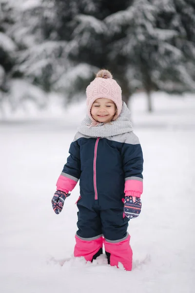 Ευτυχισμένο κοριτσάκι που περπατάει στο πάρκο με χιόνι. Αξιολάτρευτο κοριτσάκι με ροζ καπέλο και φόρμες περπατώντας στο δάσος μια χειμωνιάτικη χιονισμένη μέρα — Φωτογραφία Αρχείου