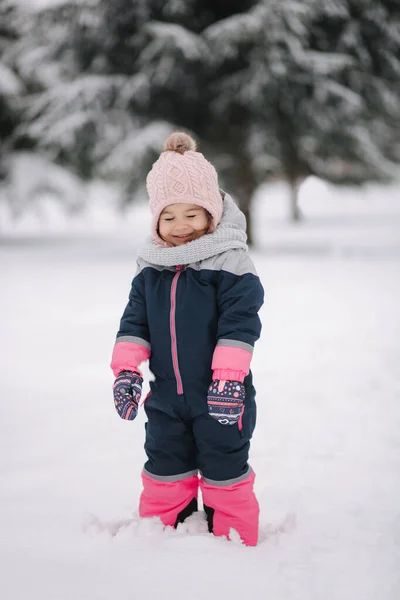 Šťastná holčička, co se prochází v parku na sněhu. Rozkošná holčička v růžovém klobouku a kombinéze procházky v lese v zimě sněžný den — Stock fotografie