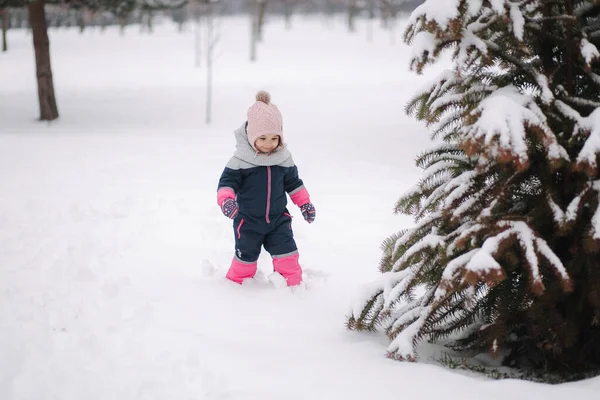 Glückliches kleines Mädchen, das im Park auf Schnee spaziert. Entzückendes kleines Mädchen mit rosa Hut und Overall, das an einem verschneiten Wintertag im Wald spaziert — Stockfoto