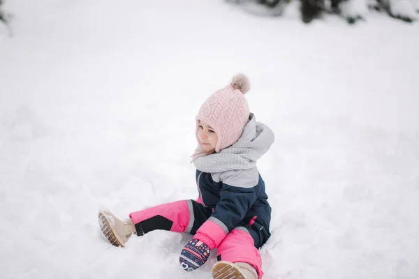 Gelukkige kleine meid die in het park op sneeuw loopt. Schattig klein meisje met een roze hoed en overall wandelend in het bos op een winterse sneeuwdag — Stockfoto